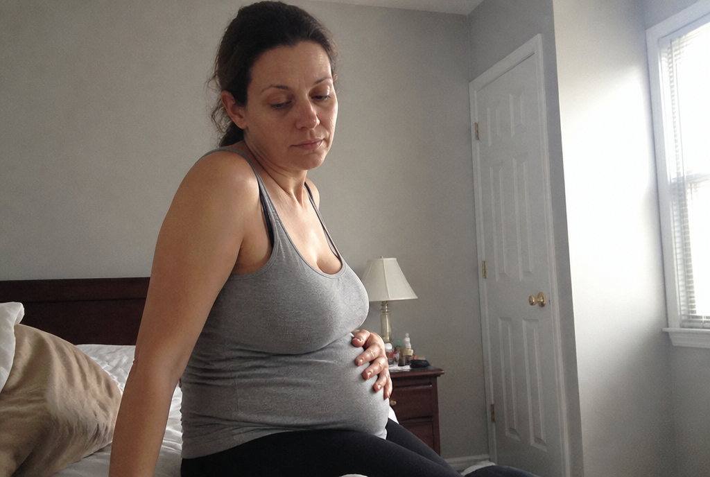 A tired woman sitting on the edge of her bed with a hand on her bloated stomach